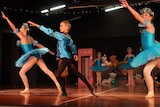 A boy in a blue ballet costumes stands between two girls on a dance stage in a ballet pose.
