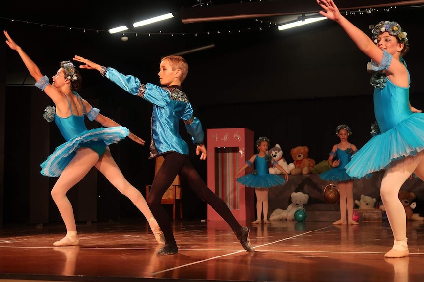 A boy in a blue ballet costumes stands between two girls on a dance stage in a ballet pose.