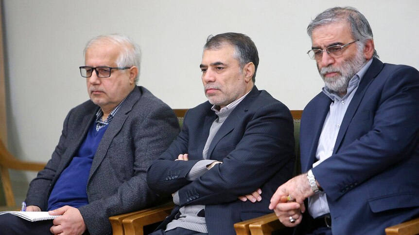 You view three greying Iranian men in navy suits sitting and taking notes on wood chairs upholstered with green cushions.