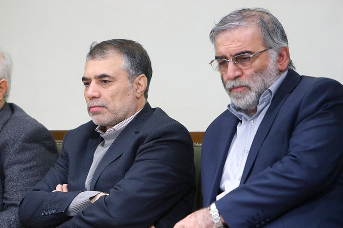 You view three greying Iranian men in navy suits sitting and taking notes on wood chairs upholstered with green cushions.