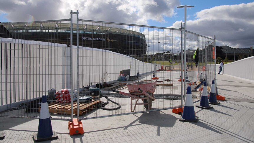 A fenced-off area at the end of the Matagarup Bridge with tools and a wheelbarrow on the ground and Perth Stadium behind.