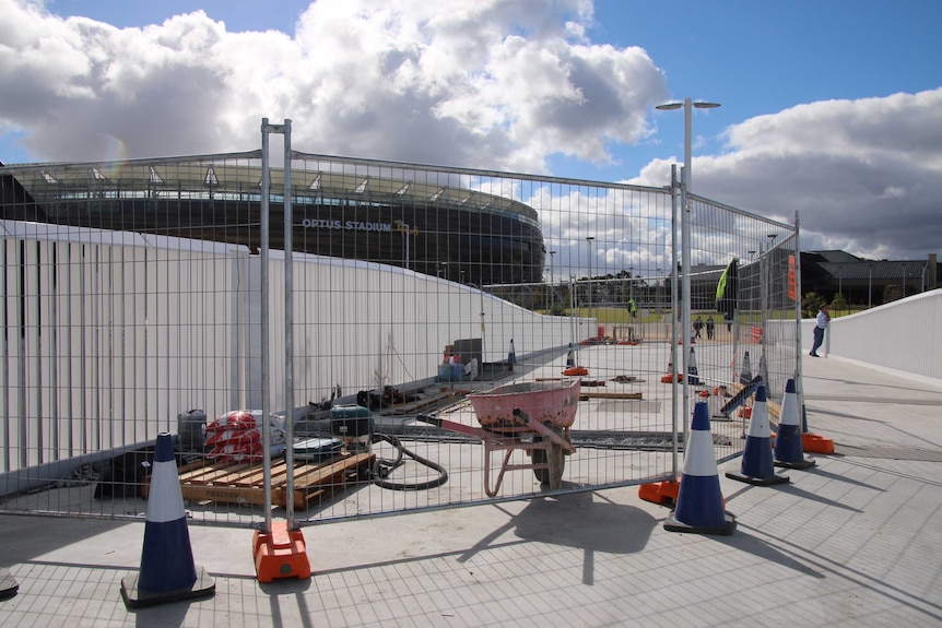 A fenced-off area at the end of the Matagarup Bridge with tools and a wheelbarrow on the ground and Perth Stadium behind.