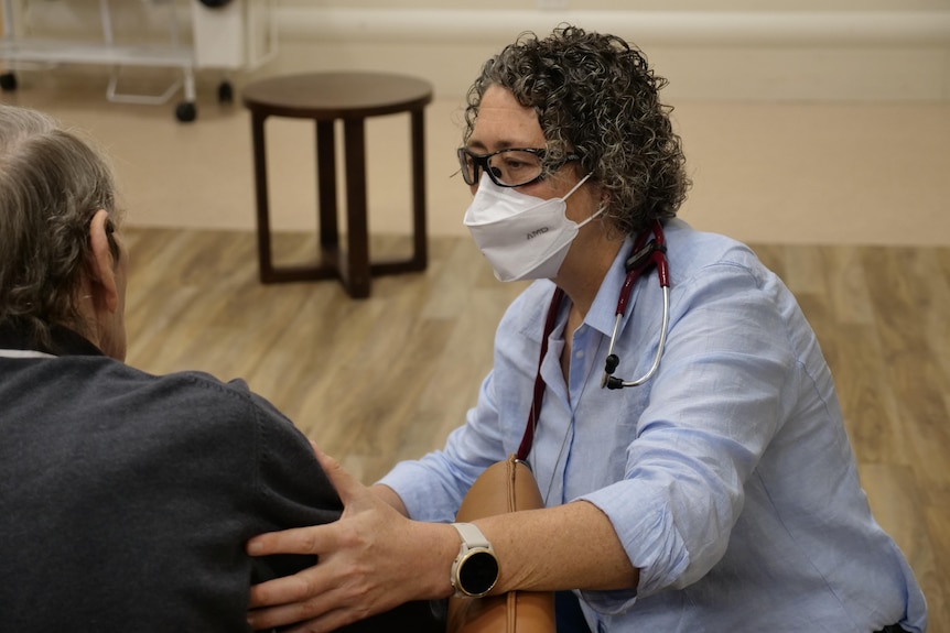 A female doctor wearing a mask, a stethoscope around her neck and glasses cares for an aged care resident