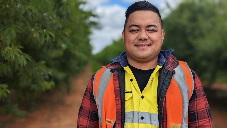 Albert Chan stands smiling among citrus trees wearing layers of black, yellow, red and orange work shirts.