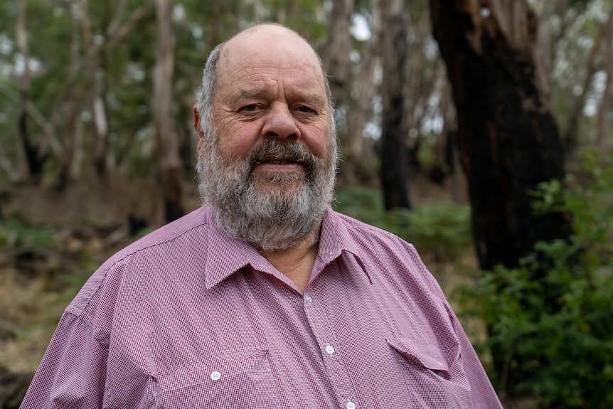 An older Indigenous man with a grey beard stands in bushland.