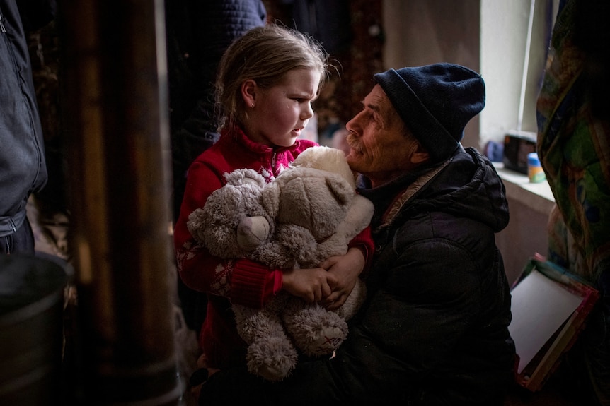 A young girl clutches to a plush toy as she sits with an older man and they look at each other.