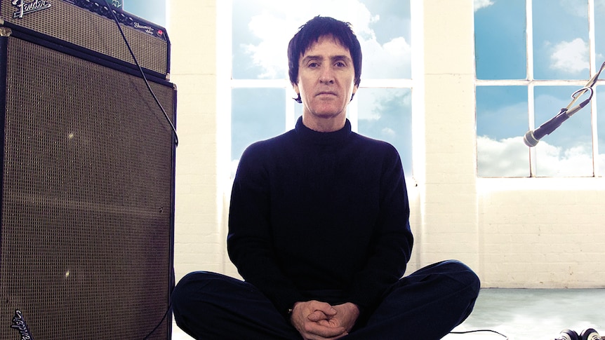 Johnny Marr wears black and sits cross-legged on the floor in front of a window. He sits between a Fender amp and a microphone.