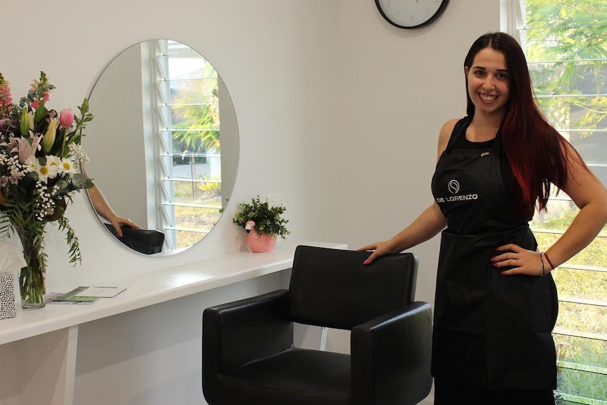 Una mujer con el pelo largo y oscuro está parado frente a la silla de un peluquero, sonriendo