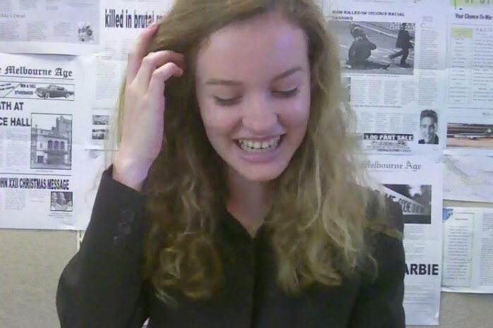 A young woman looks down smiling in an office, with newspapers on the wall in the background.