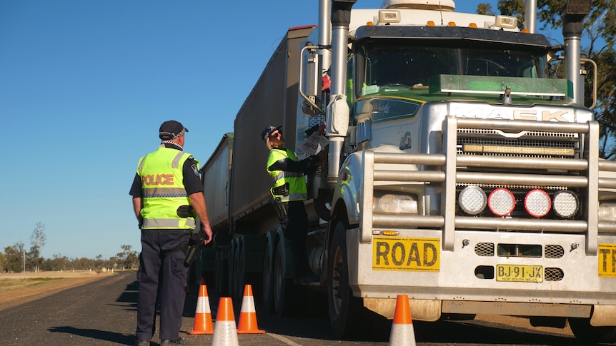 Two police officers standing next to a B-double truck. There are four orange and white cones on the road