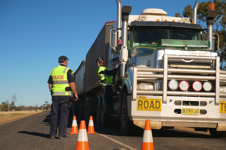 Two police officers standing next to a B-double truck. There are four orange and white cones on the road