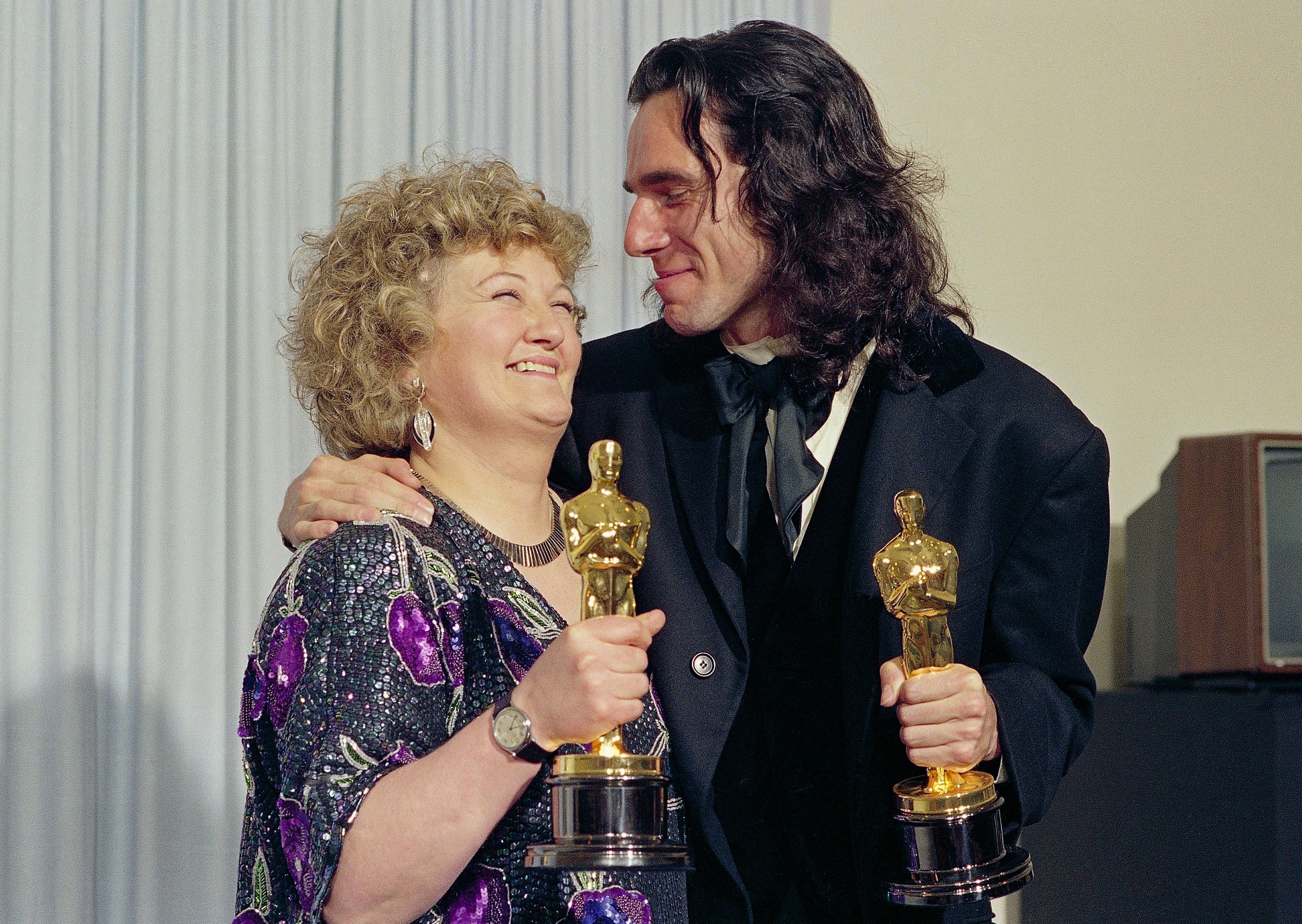 A man and a woman stand with their arms around each other holding golden trophies.