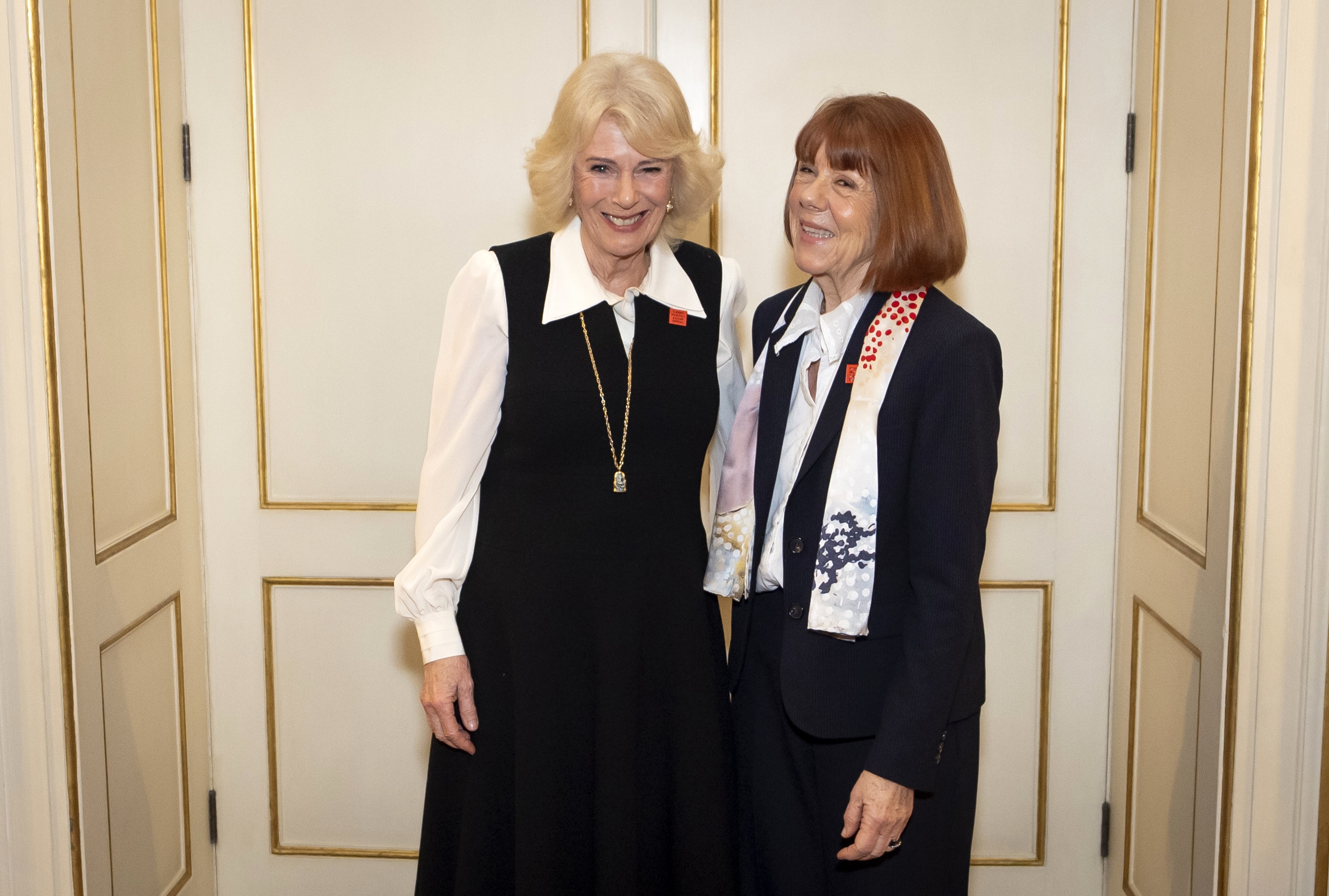 Two women, dressed in smart black and white outfits, stand side by side smiling, by cream coloured doors.