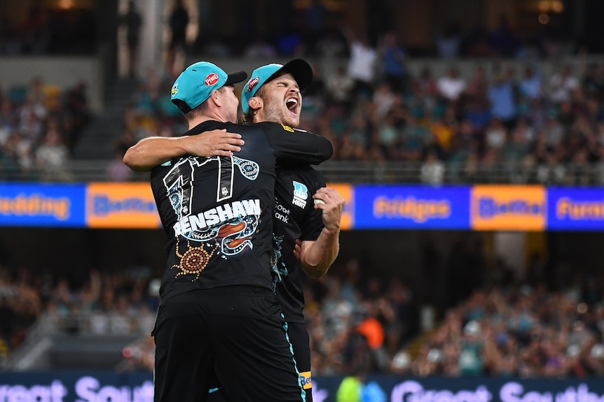 cricketers sam hain and matt renshaw hug while celebrating taking a catch wearing black uniforms and teal coloured caps