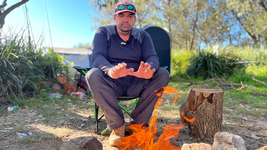 A man wearing a cap, sunglasses perched on it, sits on a folding chair by a campfire, warms his hands, stares seriously at fire.