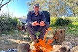 A man wearing a cap, sunglasses perched on it, sits on a folding chair by a campfire, warms his hands, stares seriously at fire.