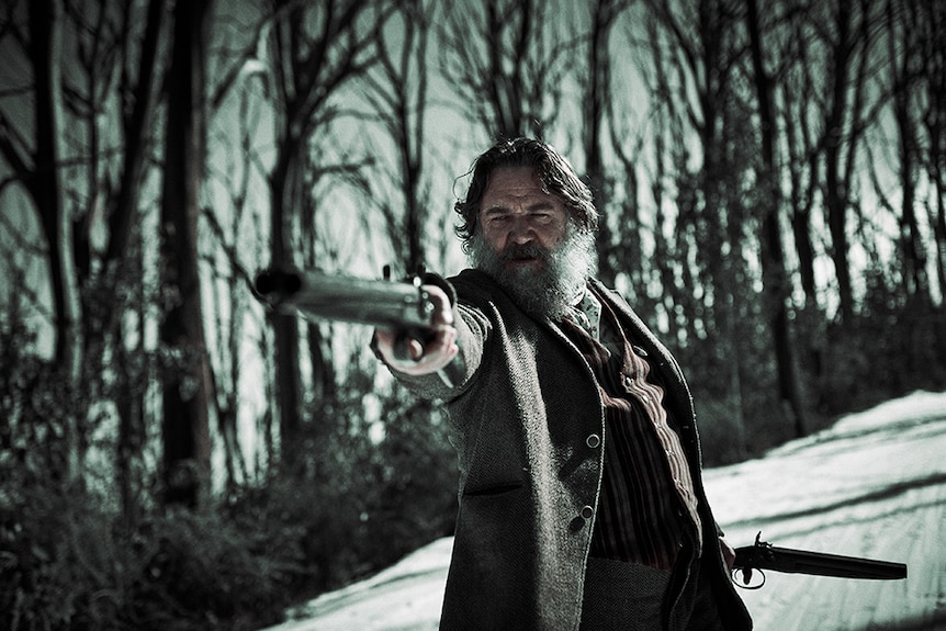 A tough looking and graying man with long beard stands with raised gun in starkly lit outback terrain amongst dead trees.