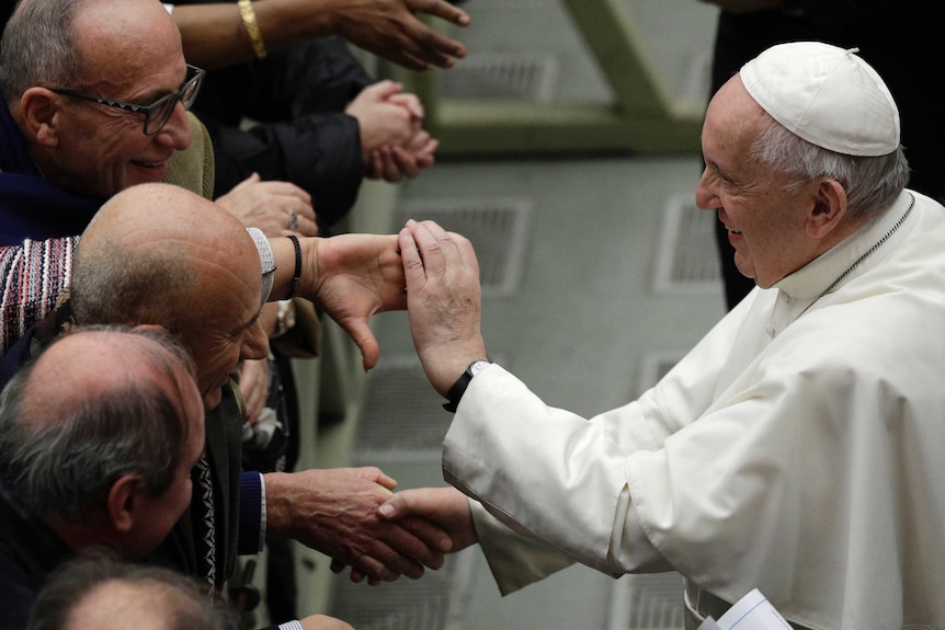 Pope in side profile of Pope in white cap shakes hands with men's outstretched hands.