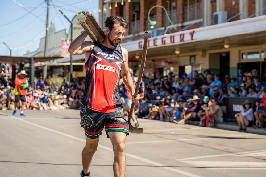 A dark-haired man in a singlet carries a load of steel down the main street of a country town.