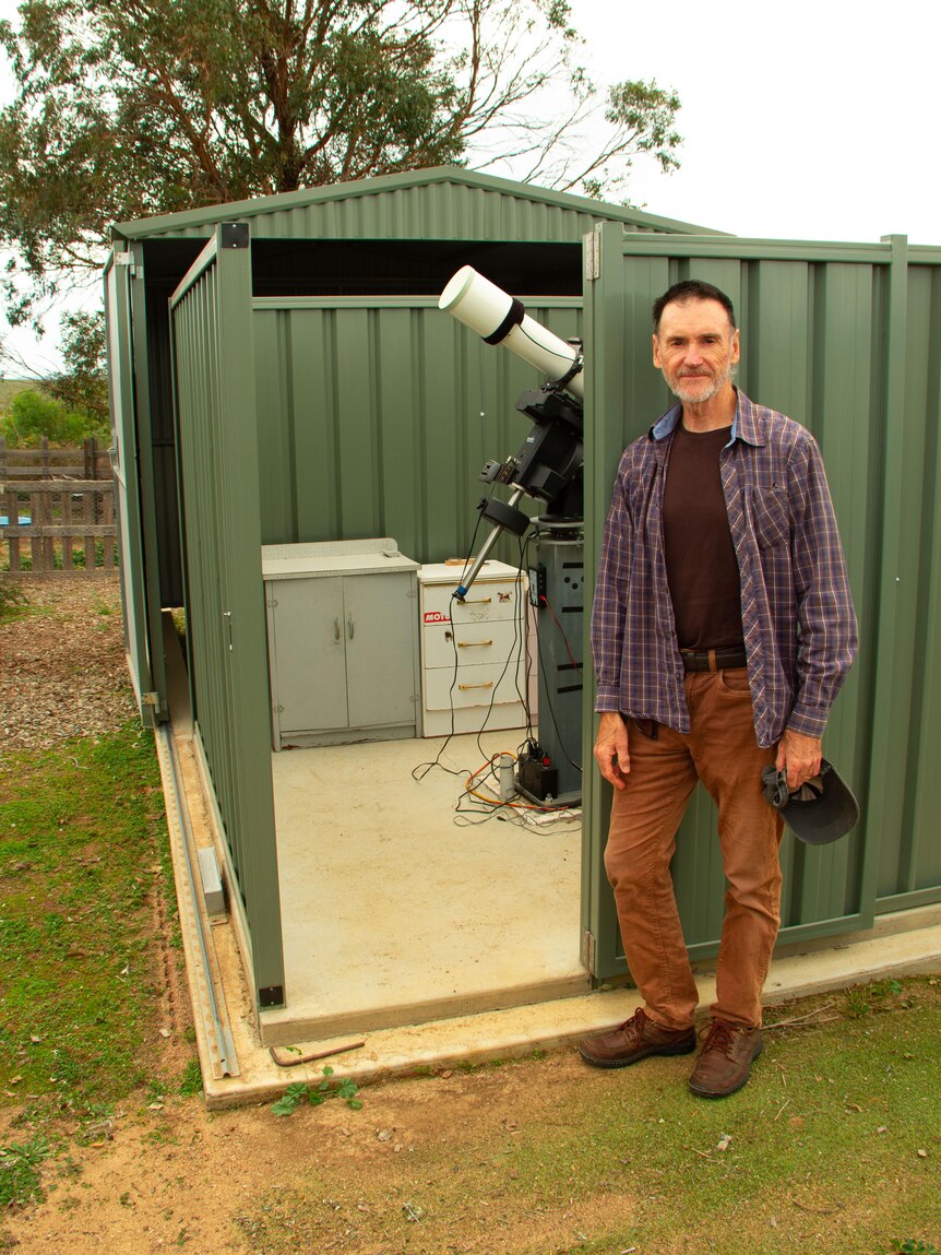 A man in a red shirt with short black hair and a grey beard stands next to a telescope in a green shed.