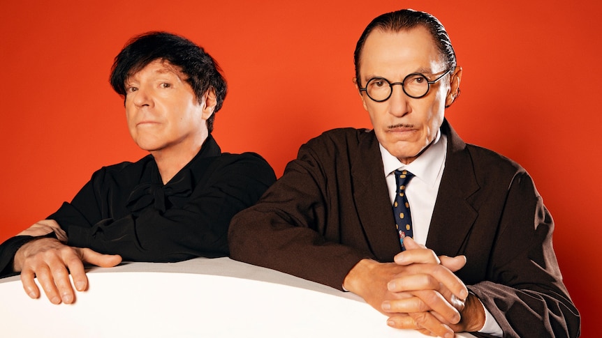 Russell and Ron Mael of Sparks stand posing on a white circle against a dark orange backdrop