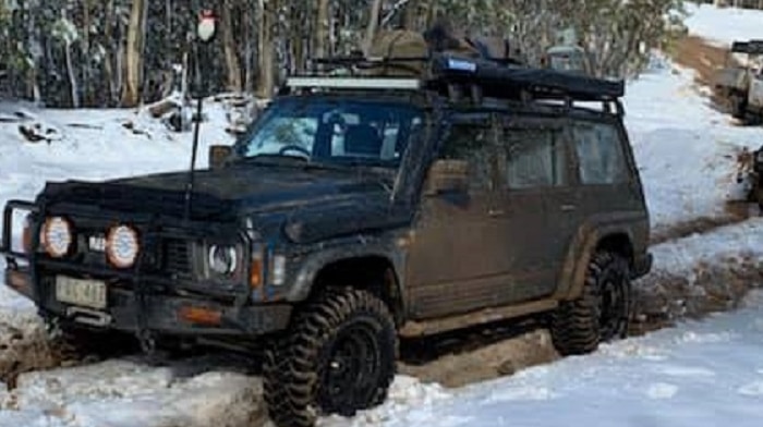 A 4WD moves along a snow-covered track in a convoy in rugged alpine terrain.
