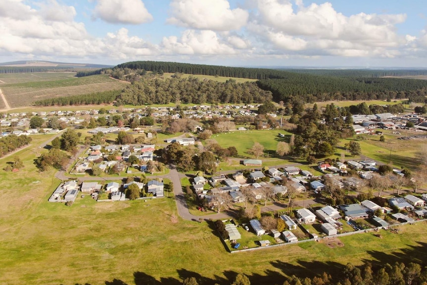 An aerial photo shows a hundred odd homes surrounded by lots of grassland and a pine forest in the distance on a sunny day.