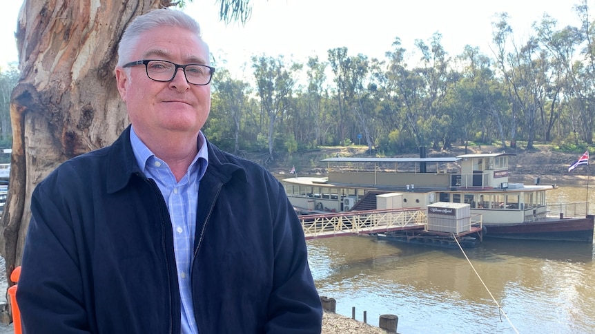 A man with grey hair and black thick-rimmed glasses stands in front of a paddle steamer docked at the riverside.