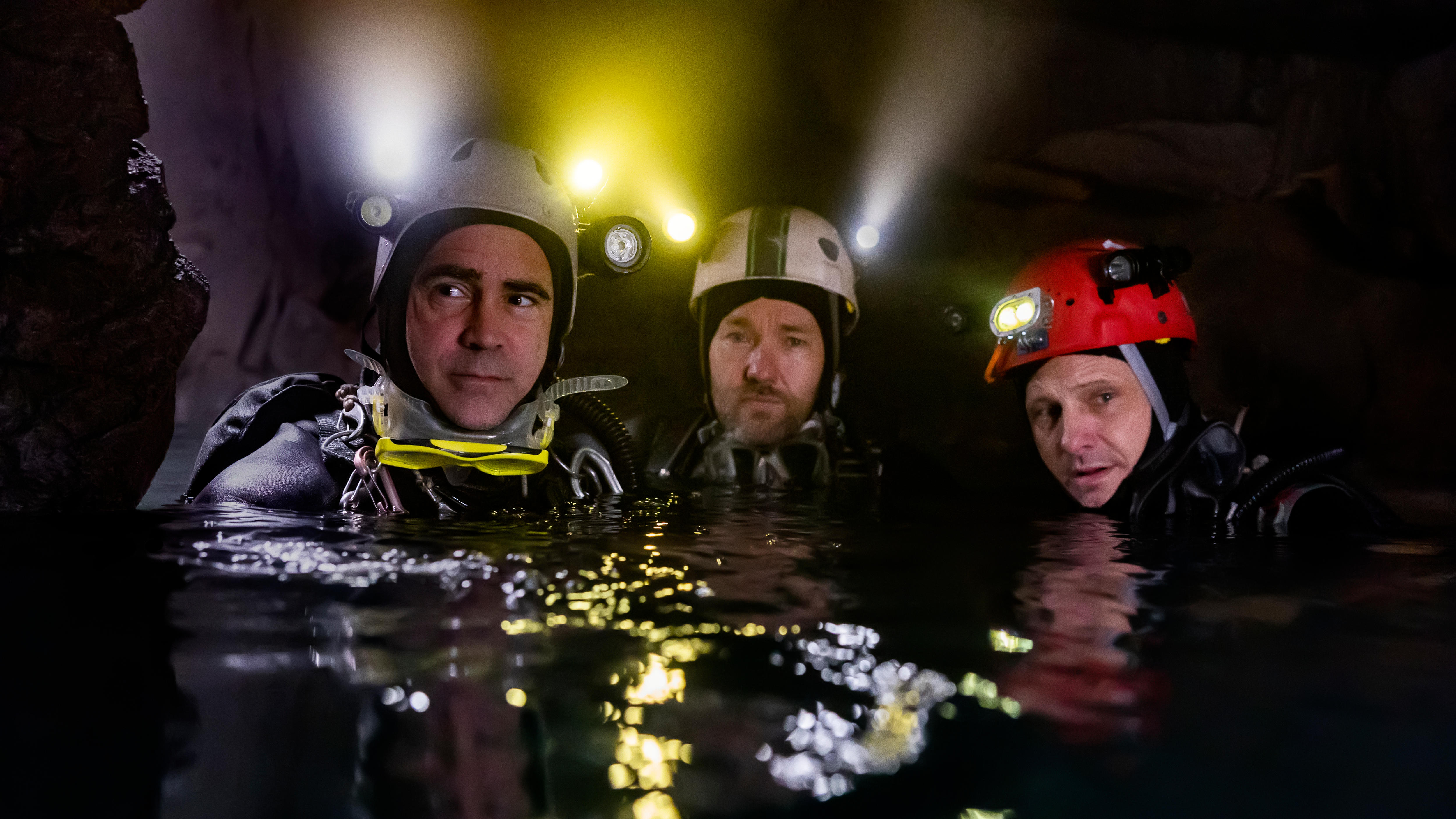 Three middle-aged white men wearing scuba-diving gear and head torches look towards the camera from chest-deep water
