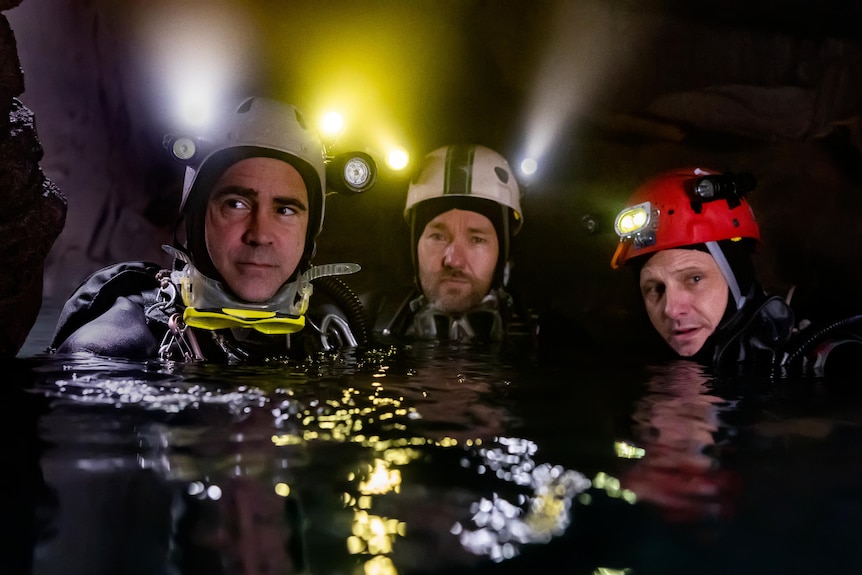 Three middle-aged white men wearing scuba-diving gear and head torches look towards the camera from chest-deep water