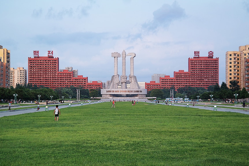 Wide colour photo of grass lawn leading up to the Monument to the Foundation of the Workers’ Party in Pyongyang with two red