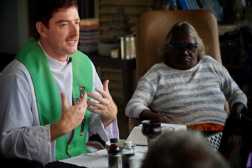 Father Pat Mara gestures with his hands and sits next to an Aboriginal woman.