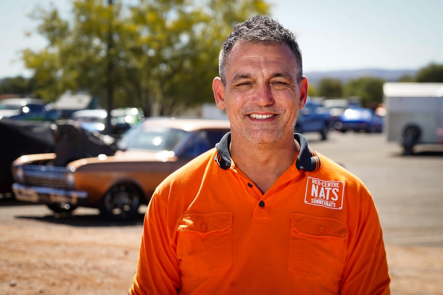 Man in bright orange shirt stands in front of some parked carss