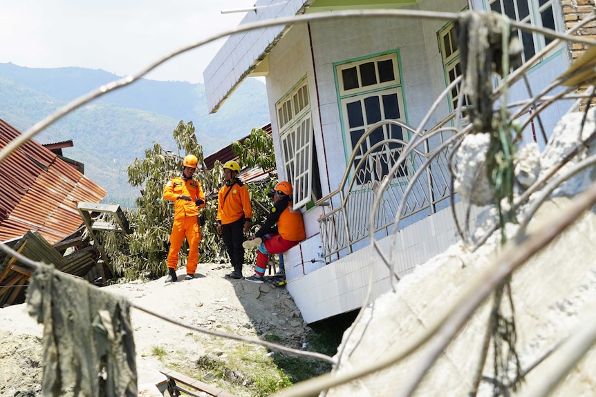 Three people in high-visibility clothes and helmets talk in the shade of a wrecked, slanting building, in mud