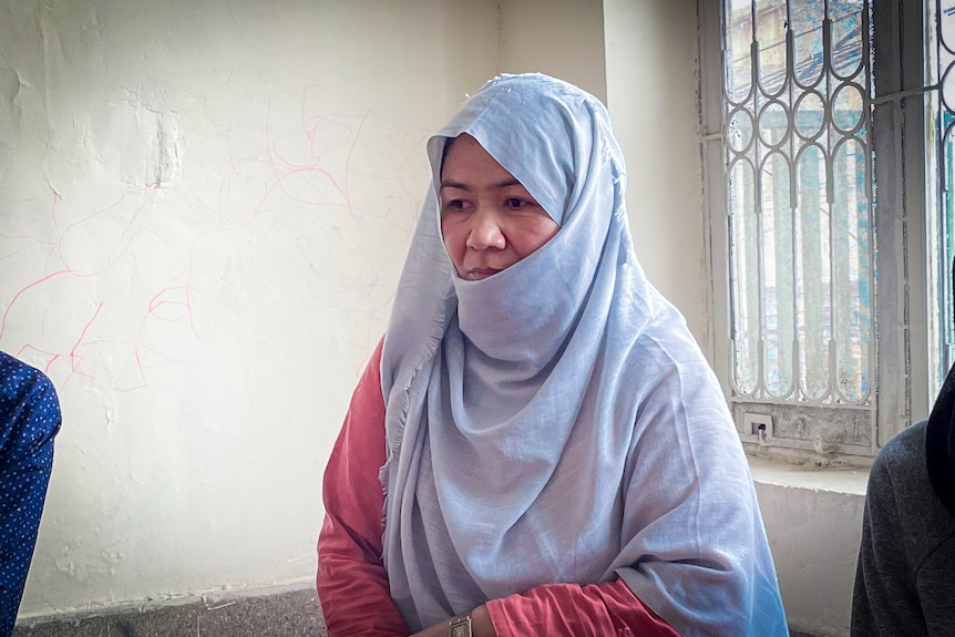 A woman in a pale blue headscarf next to a window, looking thoughtful 