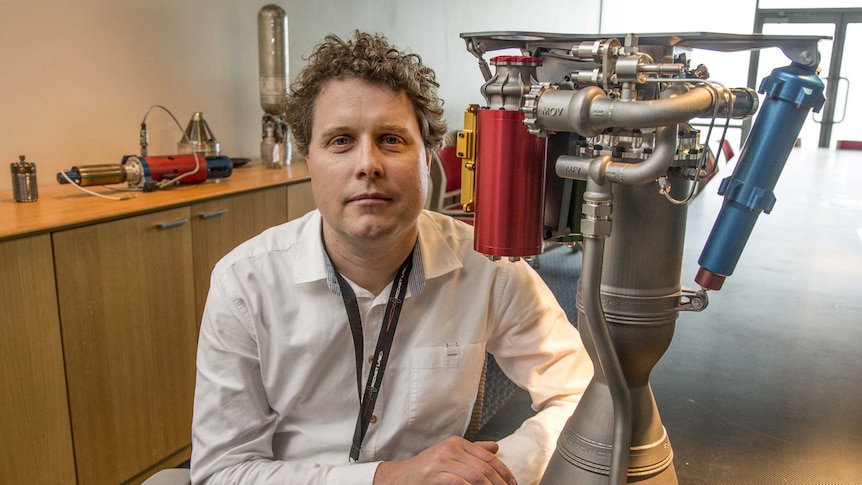 Rocket Lab CEO Peter Beck sitting next to a rocket engine on a desk.