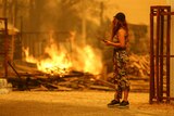 An orange-lit sky with a woman standing watching a blaze that has damaged a property. 