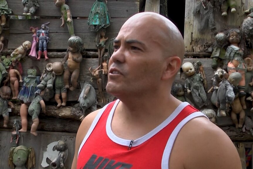 A bald man in front of a wall with dolls hanging from it