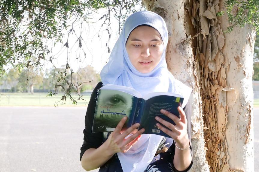 A teenager in a hijab leans against the trunk of a tree as she reads a book. The cover shows an eye and words in Arabic