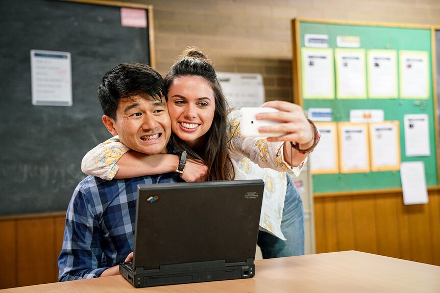A man seated at desk in a classroom winces as a smiling woman wraps arm around him and takes selfie on smartphone.