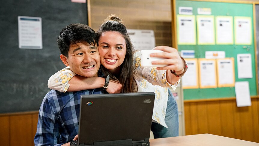 A man seated at desk in a classroom winces as a smiling woman wraps arm around him and takes selfie on smartphone.