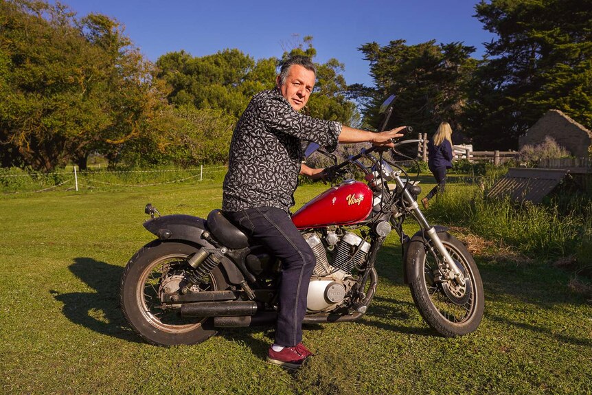 A man in a patterned black shirt and jeans sits on a red motorcycle on a farm property in the afternoon sun.