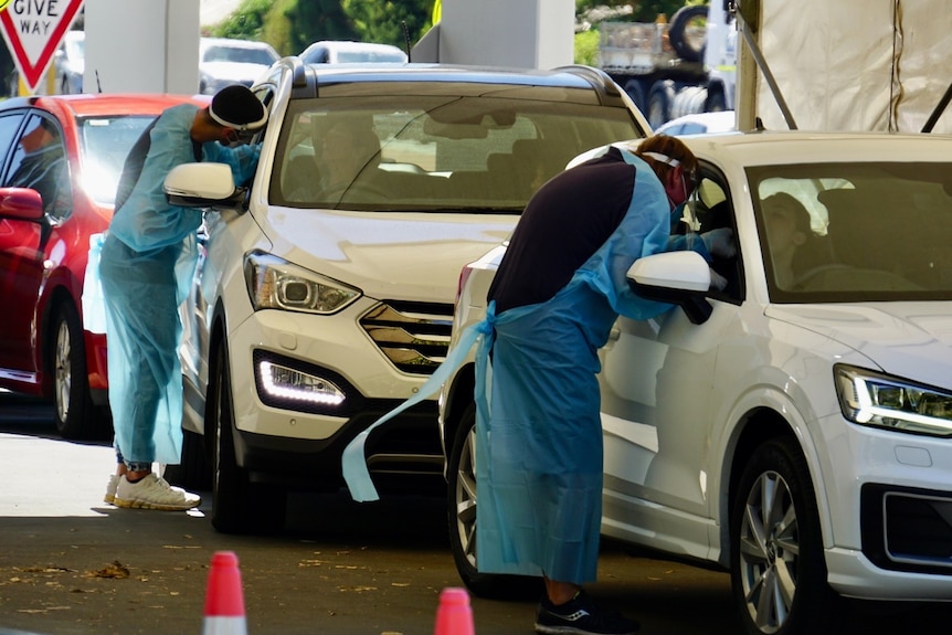 Two nurses getting a swab from drivers in two different cars in a queue at a COVID-19 testing clinic.