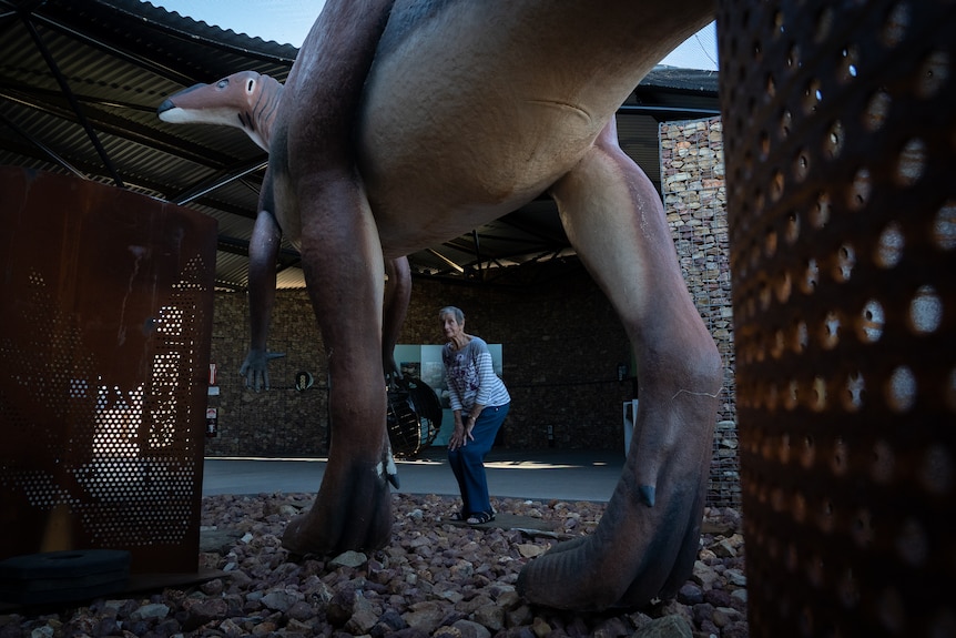 An elderly woman crouches underneath a lifesize replica of a dinosaur.