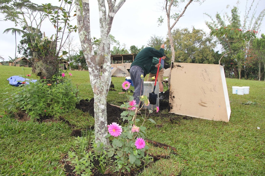 Three men dig on a grassy site near flowers next to a memorial.