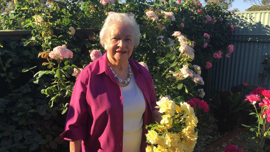 Margot White from Broken Hill stands in front of her rose bushes holding a bunch of yellow blooms.