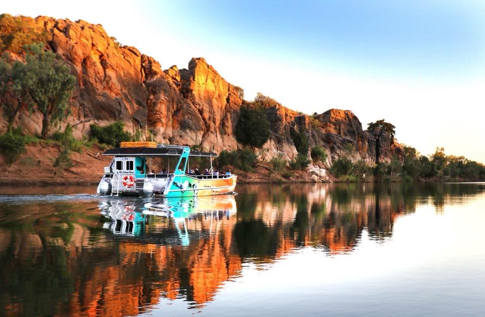 A boat cruising along the Fitzroy River, with the backdrop of red dirt cliffs and bushland