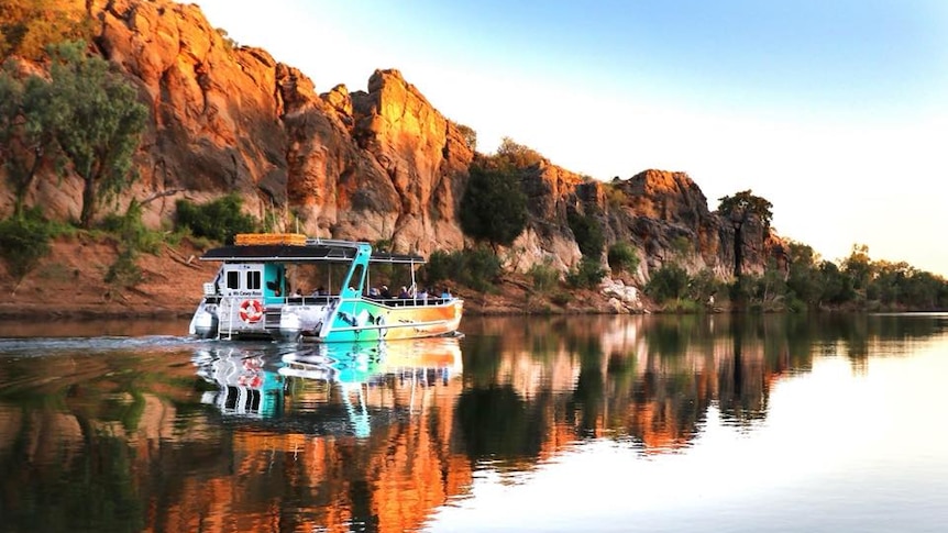 A boat cruising along the Fitzroy River, with the backdrop of red dirt cliffs and bushland