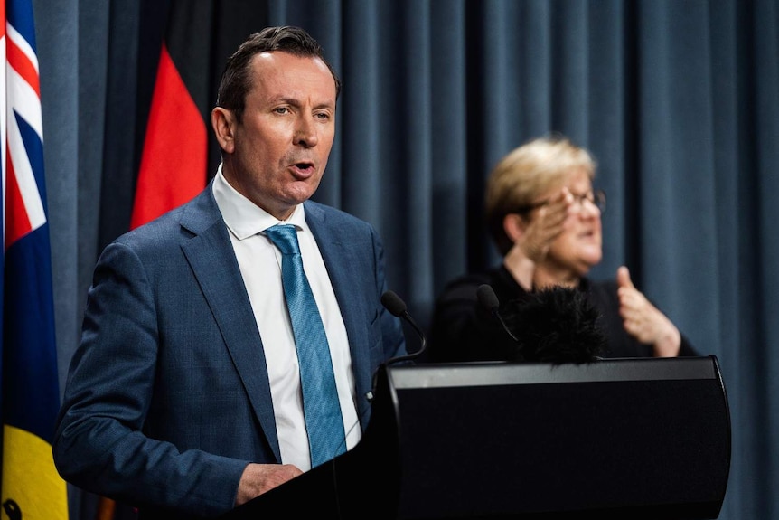 Mark McGowan wearing a blue suit, white shirt and light blue tie standing at a podium in front of a blue curtain and flags.