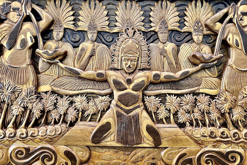 A wooden carving of a Torres Strait Islander holding two bowls and the community in the background.